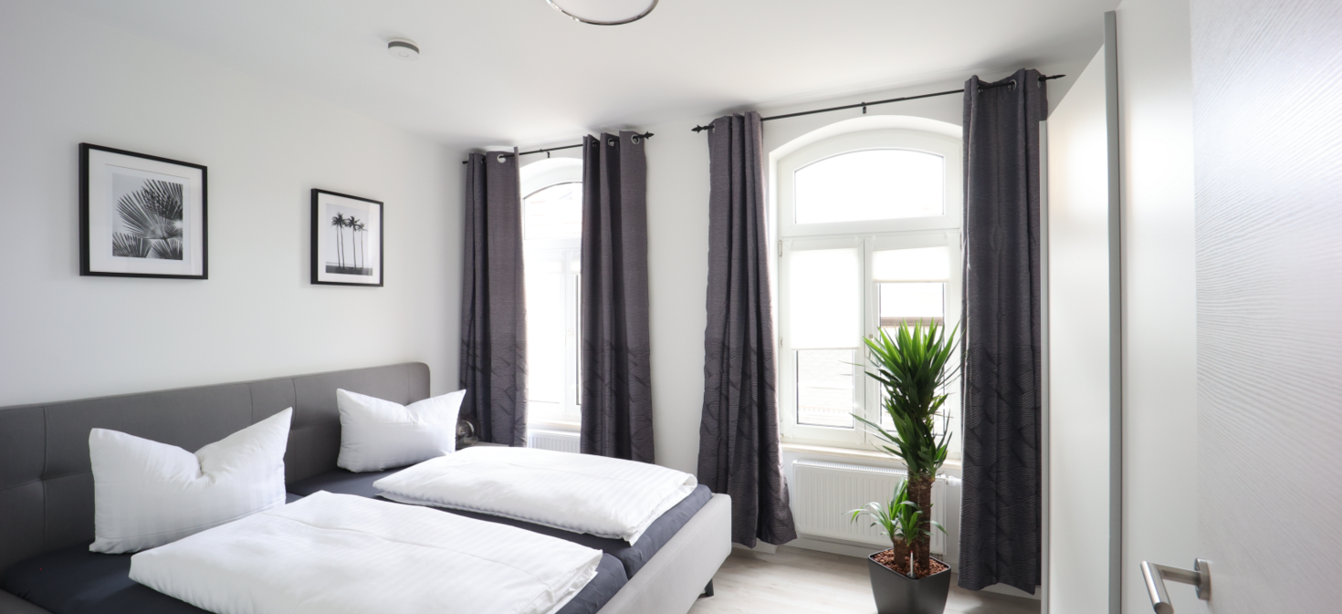 Helles Schlafzimmer mit modernem Doppelbett, Grünpflanze und großen Fenstern mit Vorhängen